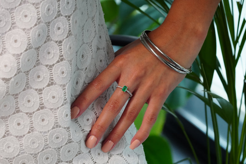 A close-up of a person's hand wearing a white gold ring with a Muzo rectangular emerald, styled with a stack of white gold bangles, next to a white lace dress and green plants.