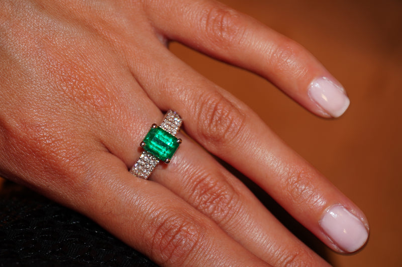 A close-up of a hand wearing a 18k White Gold ring featuring a rectangular green emerald-cut gemstone and small pavé diamonds on the band, set against a warm brown background.