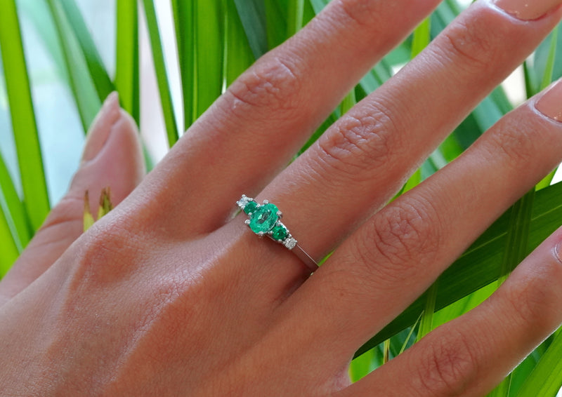 A close-up of a hand wearing a 18k White Gold ring with a central green emerald-cut gemstone and smaller side diamonds, set against a background of green palm leaves.