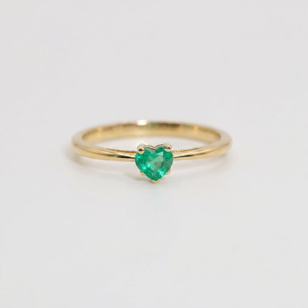 A delicate yellow gold ring featuring a small, heart-shaped green emerald gemstone in a prong setting, shown on a plain light gray background.