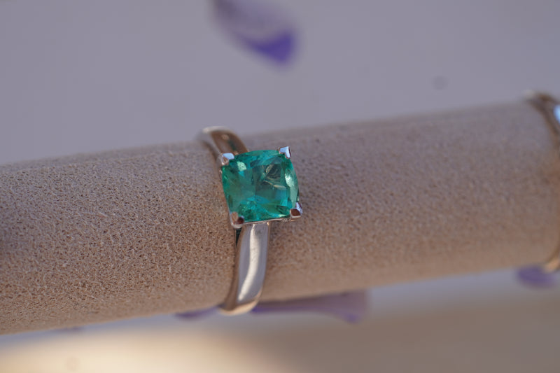 A cushion-cut emerald-cut green gemstone ring set in a 18k White Gold, displayed on a beige jewelry display stand.