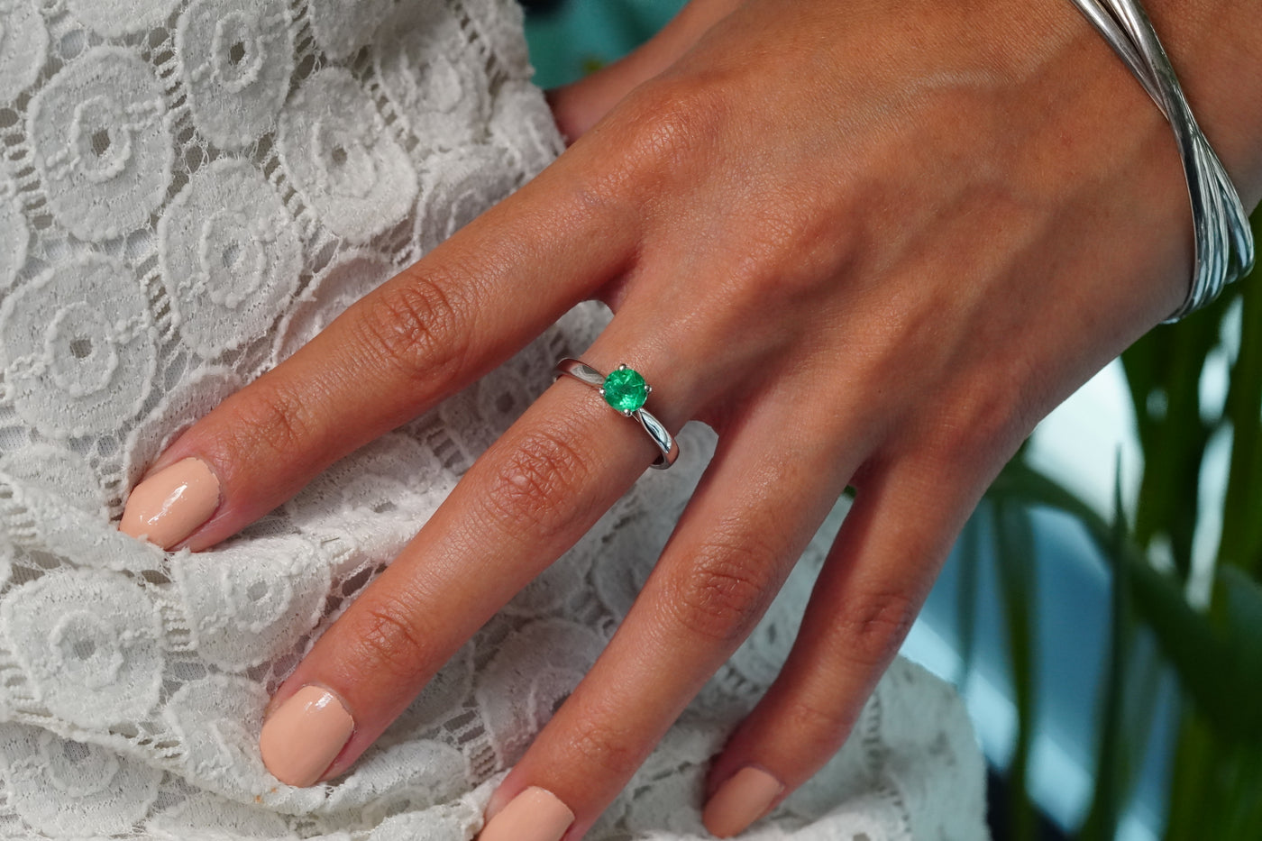 A close-up of a hand wearing a 18j White Gold ring with a round green Emerald, resting on a white lace garment.