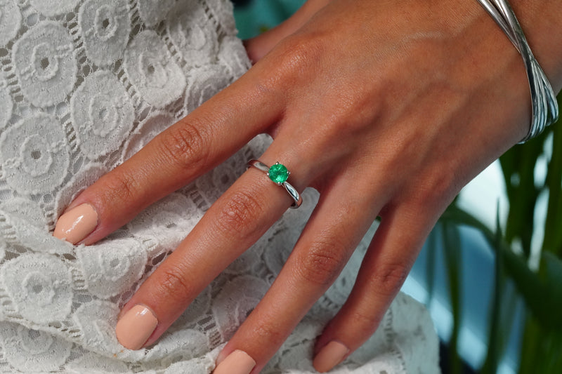 A close-up of a hand wearing a 18j White Gold ring with a round green Emerald, resting on a white lace garment.