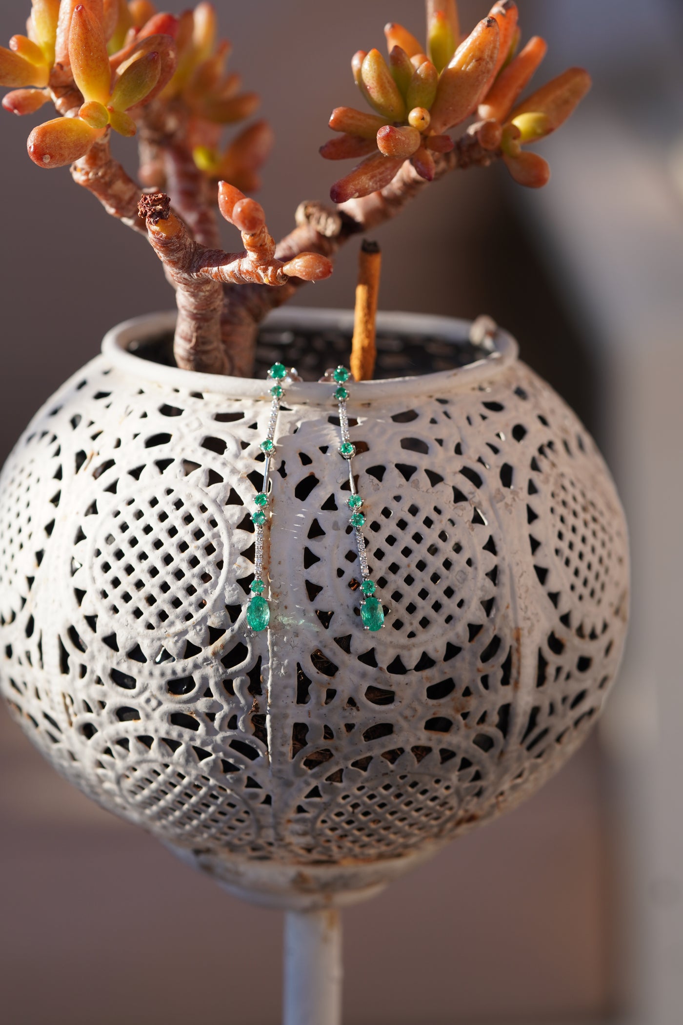 A close-up of a white spherical planter with intricate cutout lace detailing, holding a succulent plant. A pair of dangling earrings with emeralds and diamonds are draped over the front of the planter.