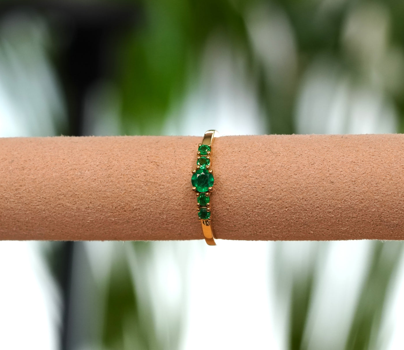 A gold band ring featuring a row of round-cut emeralds shown worn on a tan display stand with a blurred green background.