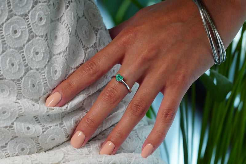 A 18k White Gold ring with a Muzo heart-shaped emerald worn on a person's ring finger, shown against a white lace dress and green plant background.