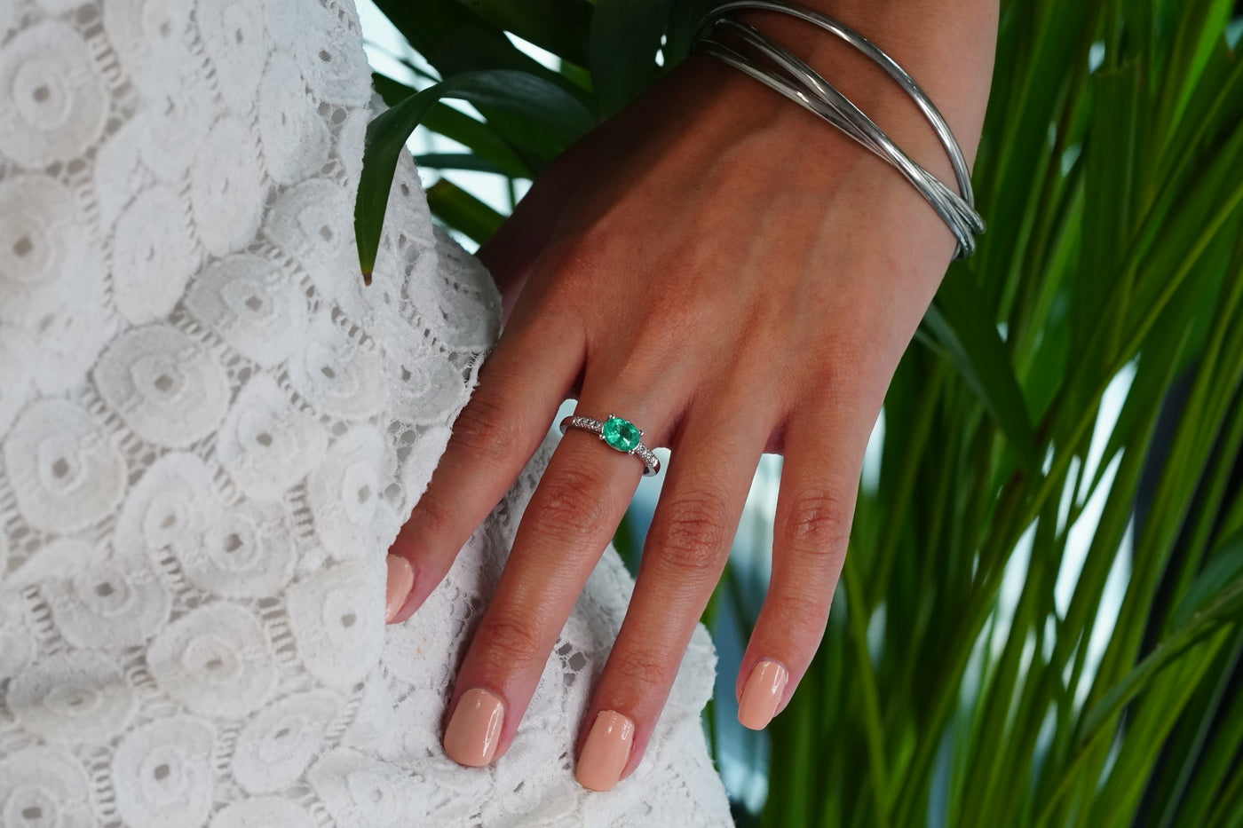 A close-up of a hand wearing a 18k white gold ring with a Muzo oval-cut emerald, resting on a white lace garment with green leaves in the background.
