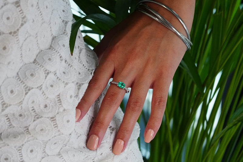 A close-up of a hand wearing a 18k white gold ring with a Muzo oval-cut emerald, resting on a white lace garment with green leaves in the background.