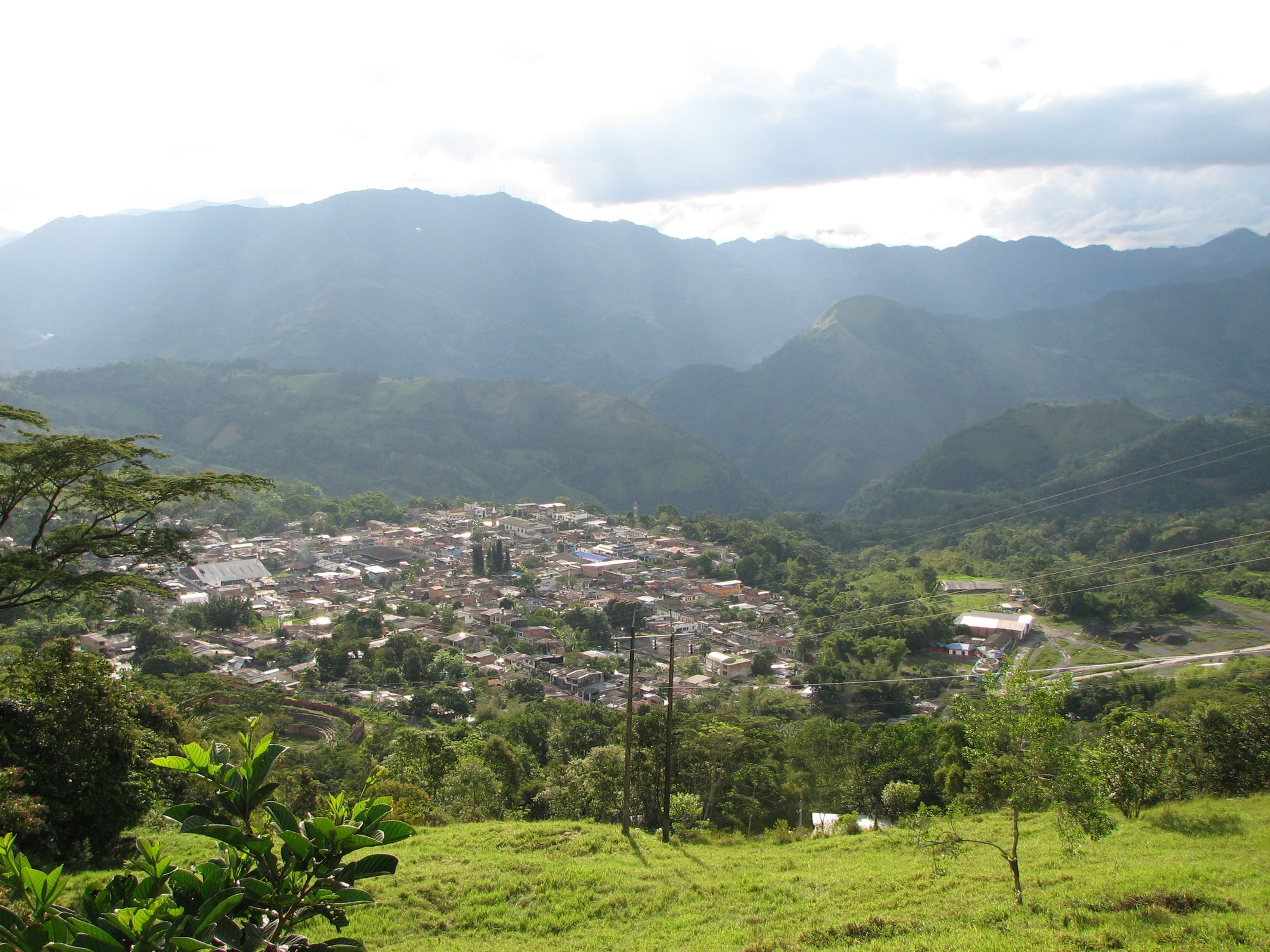A scenic landscape photograph of a small town called Muzo, nestled in a lush green mountain valley of Boyaca region in Colombia with layered blue-gray mountains in the background under a cloudy sky.