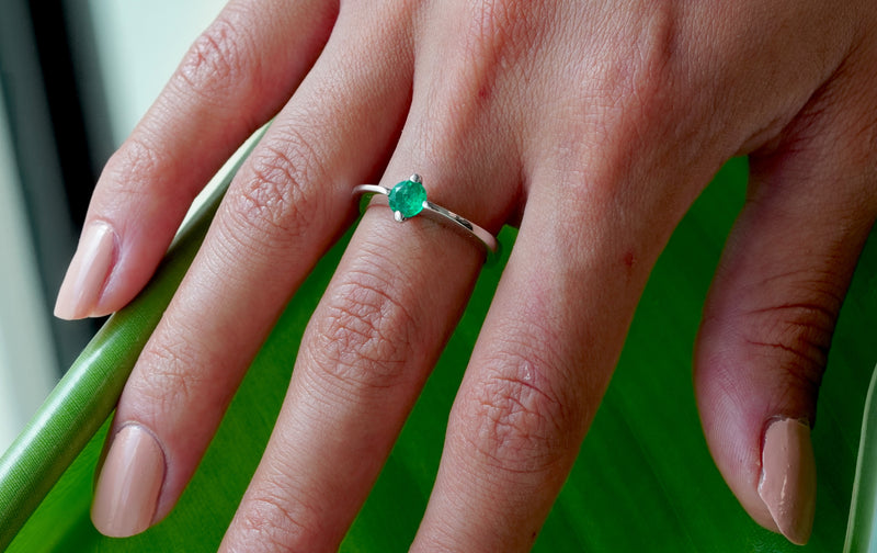 A close-up of a hand wearing a 18k White Gold ring with a round green emerald-cut gemstone, resting on a vibrant green surface.