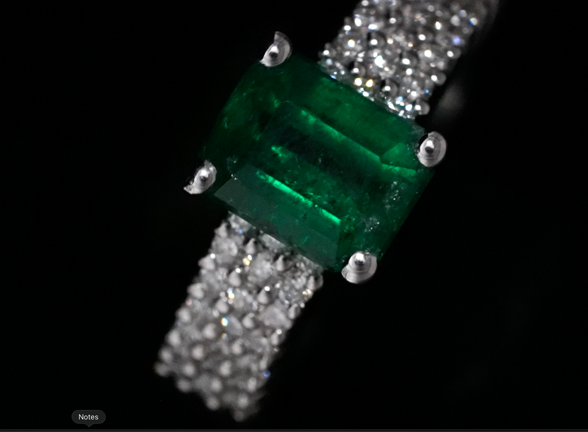 A close-up of a diamond band ring featuring an emerald-cut Muzo Emerlad in a four-prong setting, with pavé diamonds on the band, set against a solid black background.
