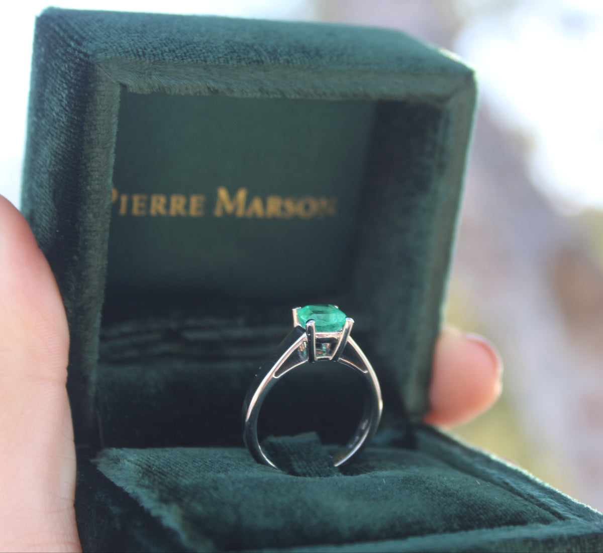 A 18k White Ring with a rectangular green emerald-cut gemstone displayed in an open dark green Pierre Marson velvet box, held by a hand against a bright, blurred outdoor background.